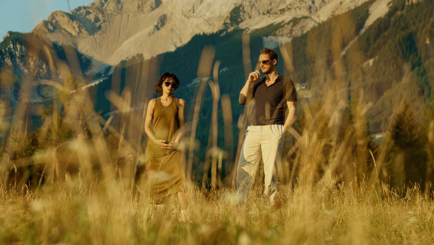 Femme en robe olive et homme en polo sombre portant des lunettes de soleil Rolf dans une prairie de montagne